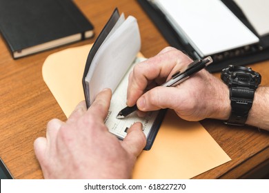 Man With Watch Writing A Check In A Checkbook. Envelope And Notepads Are Nearby.