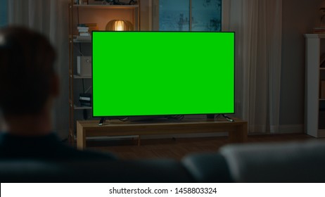 Man Watches Green Mock-up Screen TV While Sitting On A Couch At Home In The Evening. Cozy Living Room With Warm Lights.