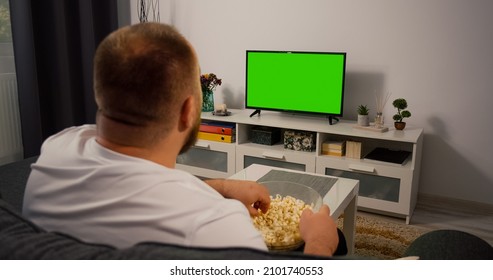 Man Watches Green Mock-up Screen TV While Sitting On A Couch At Home In The Evening In Living Room Over The Shoulder Shot.