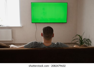 A Man Watches A Large TV Sitting On The Couch. Rear View. A Man Is Watching TV Hanging On The Wall. Green Screen TV.