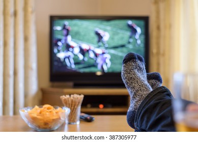 Man Watching American Football Match On TV (television) With Feet On Table, Eating Snacks - Stock Photo