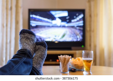 Man Watching American Football Match On TV (television) With Feet On Table, Eating Snacks - Stock Photo