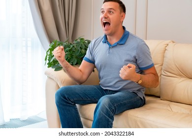 Man Watching Football In Living Room. Millenial Man At Home Alone Watching Soccer Games On Tv Enjoying And Celebrating Goal And Gestures Of Victory Sitting On Couch Happy And Excited