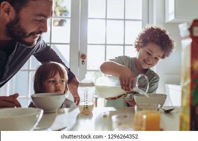 Man Watching His Kid Pour Milk In His Breakfast Bowl. Father And Kids Sitting At The Table Preparing Breakfast.