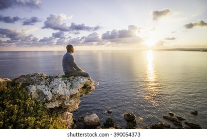 Mann beobachtet Sonnenaufgang, der auf Felsen neben dem Kliff sitzt. Malta, Europa