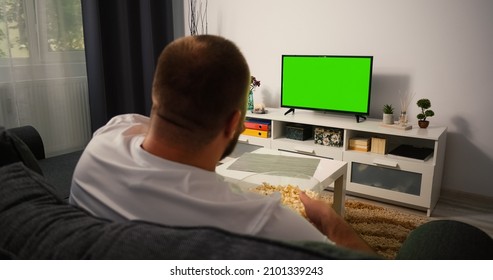 Man Watching Television With Green Screen Chroma Key, Shot Behind Models Shoulders. Man Eating Popcorn Relaxing Sitting On A Couch Home Looking To TV.