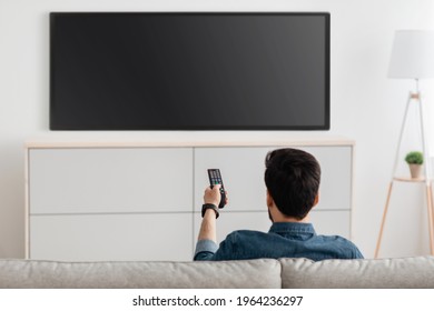 Man Watching Television, Turning On Plasma Flatscreen TV-set, Pointing Remote Control At Empty TV Screen On White Wall. Guy Switching Channels At Home, Back View. Mockup