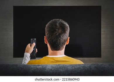 Man Watching Television, Turning On Plasma Flatscreen TV-set, Pointing Remote Control At Empty TV Screen On Wall. Guy Switching Channels At Home, Back View. Mockup