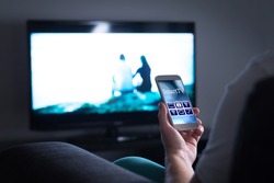 Man Watching Television And Using Smart Tv Remote Control Application On Mobile Phone. Choosing Movie Stream, Switching Channel Or Changing Settings In The Menu And User Interface With Smartphone.