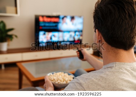 Man watching tv and eating popcorn at home on a couch, back view