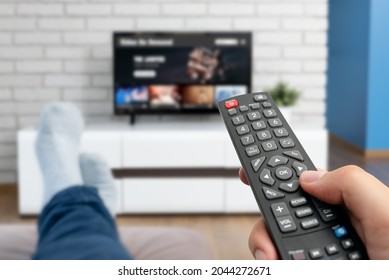 Man Watching TV, Lying On Sofa, Legs On Table. Person Holding Remote Control In Living Room