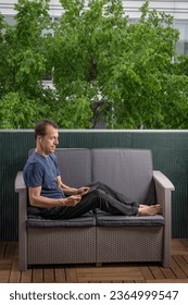 A man watching working with a digital tablet and headphones on the head, sitting on a sofa on a balcony, with trees in the background - Shutterstock ID 2364999547