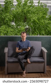 A man watching working with a digital tablet and headphones on the head, sitting on a sofa on a balcony, with trees in the background - Shutterstock ID 2365098727