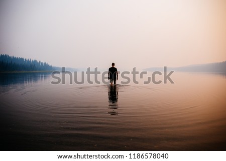 Man in the water knee deep. Smokey atmosphere. Spooky. Reflection in the water. Mountains and trees in the background.