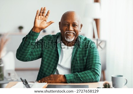 Man Waves Hello While Sitting at a Desk in a Bright Room During the Day