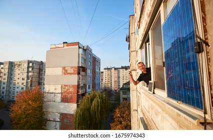 Man waves on the balcony of a multi-storey building equipped with a solar panel, against a background of a residential area of the city with clear blue sky in the warm sunny evening