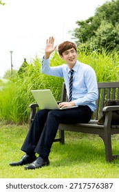 Man waves while using laptop on bench in park during sunny day