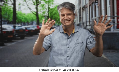 Man waving both hands and smiling with visible palms on a city street with parked cars, trees and bicycles; greeting happiness.