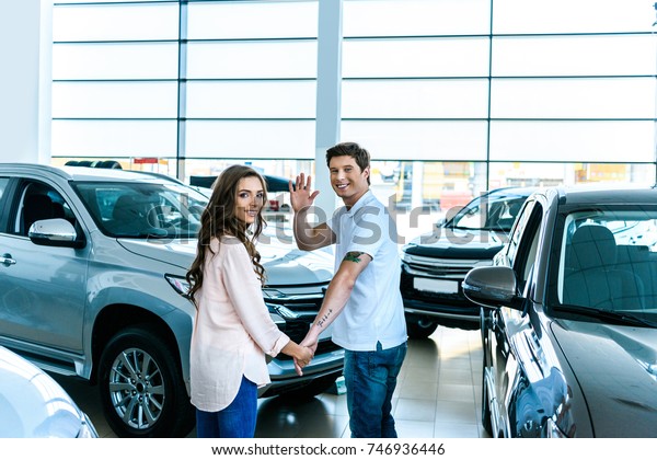 Man waving a hand and holding hands with his\
girlfriend in a car\
showroom