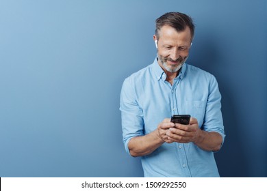 Man Wearing Audio Ear Buds Holding A Mobile Phone And Smiling As He Reads Something On The Screen Against A Blue Studio Background With Copy Space