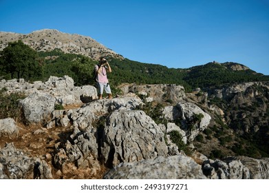 Man wearing backpack and using binoculars while exploring rocky mountain terrain. The scene depicts adventure, exploration, and nature appreciation under a clear blue sky. - Powered by Shutterstock