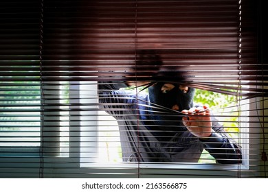  Man Wearing Balaclava Face Mask Looking Through Venetian Blinds. Keep Your Home Secure During Your Summer Holiday Concept.