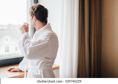 Man wearing bathrobe drinking coffee in front of a window - Powered by Shutterstock