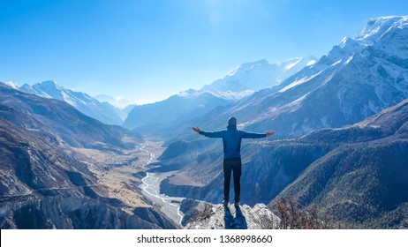 Man wearing a beanie and blue jumper, spreads his arms wide, breathing deeply the fresh mountain air. His gesture represents freedom and happiness. Below a long valley stretches in Himalayas.