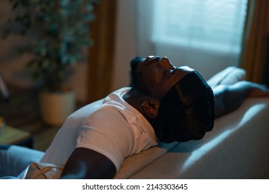 Man wearing black bandana is resting on couch with head tilted back. Men with stubble is experiencing negative emotions, breakdown, sad day, dark night lantern light from outside window.