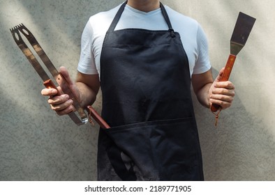 A man wearing black chef's apron, holding barbecue tools: bbq tongs, spatula. - Shutterstock ID 2189771905