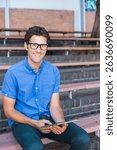 Man wearing black glasses and blue shirt sitting on schoolyard bleachers holding tablet. Outdoor seating, casualwear, educational, technology, leisure, rustic, urban