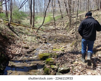 Man wearing black hat and coat, blue jeans and muddy boots strolling along shallow creek bed lined in mossy rocks running through late fall or early spring forest with light green leaves and blue sky. - Powered by Shutterstock