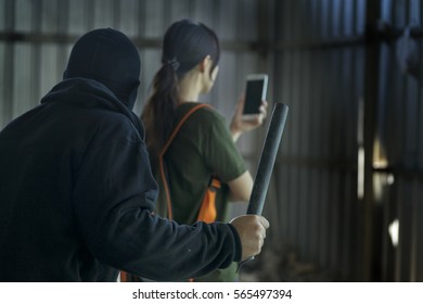 Man Wearing A Black Hood Mask Holding A Rod Of Iron To Hurt The Girl And Standing Behind, Woman Playing A Mobile With No Careful - Dark Tone - Un-safety Concept