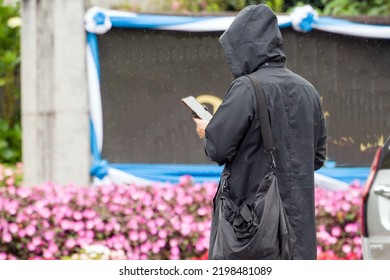 A Man Wearing A Black Raincoat Stands Looking At Smartphone  In The Middle Of A Small Rain.