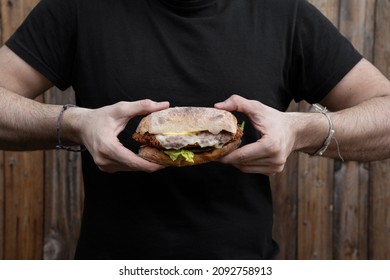 Man Wearing A Black Shirt, Holding A Chicken Fried Steak Sandwich With Lettuce And Mayonnaise. 