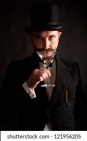 Man wearing black suit and top hat with a raised martini glass to his lips about to drink it. This image was shot with one light to give it drama and a grungy appearance