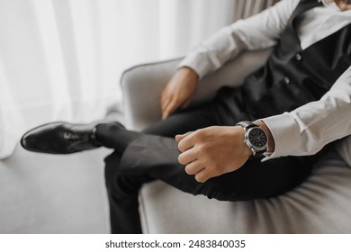 A man wearing a black vest and a white shirt is sitting on a couch with his hand on his wrist. He is wearing a watch and he is relaxed - Powered by Shutterstock