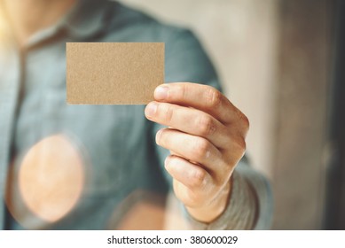 Man wearing blue jeans shirt and showing blank craft business card. Blurred background. Horizontal mockup