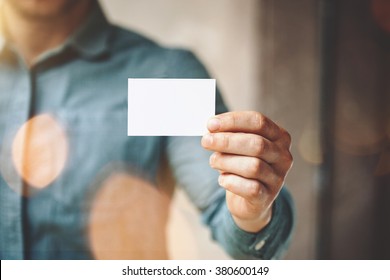 Man wearing blue jeans shirt and showing blank white business card. Blurred background. Horizontal mockup
