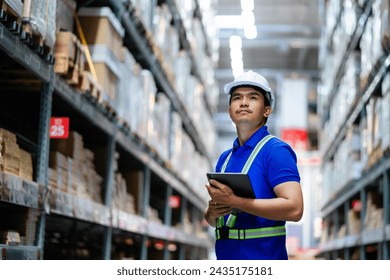 A man wearing a blue shirt and a hard hat is standing in a warehouse with a tablet in his hand. - Powered by Shutterstock