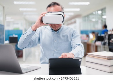 A man wearing a blue shirt is wearing a virtual reality headset and playing a game on a tablet. Seniors in technology classes with laptops, tablets, VR headsets. Adult learning - Powered by Shutterstock