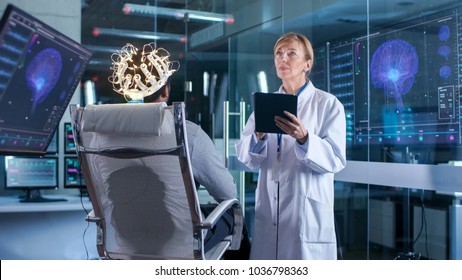 Man Wearing Brainwave Scanning Headset Sits in a Chair while Scientist with Tablet Computer Supervises Process. In the Modern Brain Study Laboratory Monitors Show EEG Reading and Brain Model.