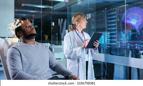 Man Wearing Brainwave Scanning Headset Sits in a Chair while Scientist with Tablet Computer Supervises Process. In the Modern Brain Study Laboratory Monitors Show EEG Reading and Brain Model.