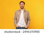 A man wearing a brown button-up shirt and jeans stands against a yellow background. He is smiling directly at the camera with his hands in his pockets.