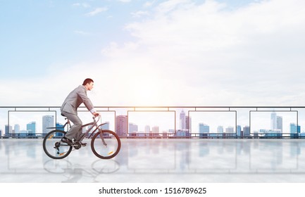 Man wearing business suit riding bicycle on penthouse balcony. Handsome cyclist on background of city center architecture. Terrace with modern downtown view. Real estate agency business concept