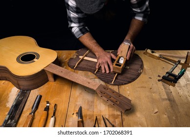 Man wearing cap and plaid shirt making guitar. Planning guitar back. Luthier's workshop. Dark black background.