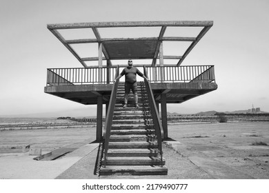 A Man Wearing Casual Clothes, Stylish Sunglasses And Standing In An Empty Abandoned Pool And Holds On To The Stairs. Diving Board In The Outdoor Pool