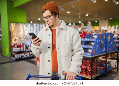 Man wearing checks his phone while pushing shopping cart in store decorated with holiday items. Pre-Christmas gift shopping at the supermarket. - Powered by Shutterstock