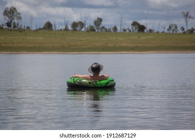 Man Wearing A Cowboy Hat, Floating In A Rubber Ring On A Lake, Watching The Approaching Storm.