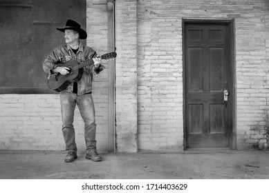 Man wearing cowboy hat and jeans, playing acoustic guitar alone outside on old dirty building porch, Spanish classical guitar, singing, performing - Powered by Shutterstock